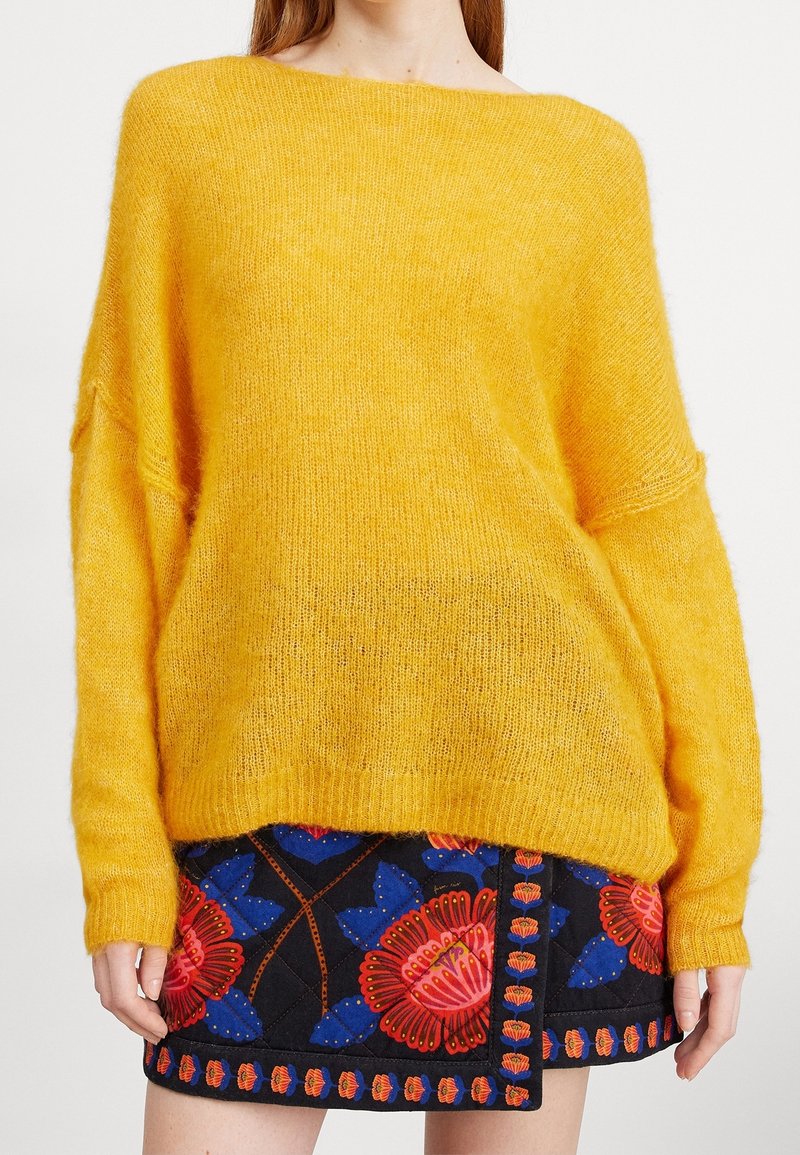 Gul överstor tröja med en mjuk, fluffig textur, båthals, och ribbade ärmslut, kombinerad med en svart blommönstrad kjol.