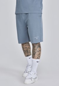 Șorturi din bumbac de culoare albastru deschis, cu buzunare laterale, având un logo SIKSilk în alb. Împreună cu șosete albe și adidași.