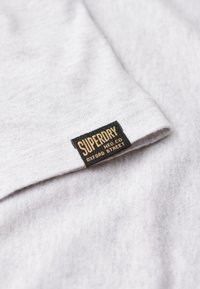 Lys grå bomuldsstof med en blød tekstur, med et sort label med "SUPERDRY MFG CO OXFORD STREET" i guld skrifttype.