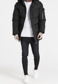 Chaqueta acolchada negra con capucha forrada de piel, múltiples bolsillos y un bolsillo con cremallera en el brazo, combinada con una camisa blanca y vaqueros ajustados negros.