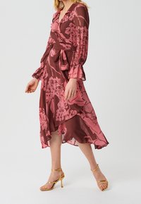 Vestido envolvente floral rosa con mangas largas, puños con volantes y un dobladillo asimétrico. Fabricado en un tejido ligero y transparente. Tacones dorados con tiras.