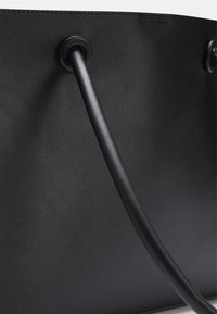 Musta nahkatasku, jossa on teksturoitu pinta, varustettuna sileällä mustalla kahvalla, joka on kiinnitetty pyöreällä metalliosalla, ja minimalistinen muotoilu.