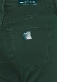 Gröna denimjeans med en närbild av bakfickan, som har en etikett märkt "A|X" och subtila sömnadsdetaljer.