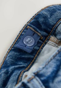 Detail tkaniny denim s modrým knoflíkem, kontrastním hnědým prošíváním a bílou vnitřní podšívkou. Texturovaný povrch s mírným leskem.