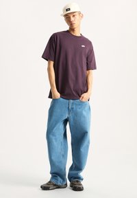 Camiseta de algodón morada con un logo, combinada con jeans de mezclilla azul holgados y zapatillas grises. El modelo lleva una gorra color crema. Fondo liso.