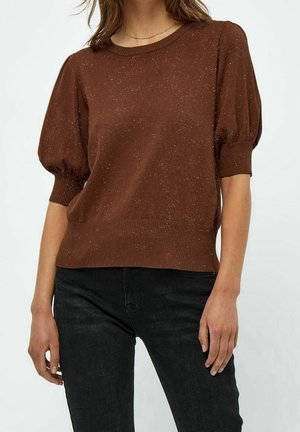 Femme portant un pull court à manches bouffantes marron tacheté, associé à un jean noir, debout devant un fond clair uni.