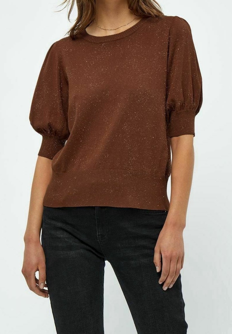 Femme portant un pull court à manches bouffantes marron tacheté, associé à un jean noir, debout devant un fond clair uni.