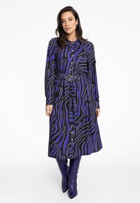 Langärmliges Shirtkleid aus gemustertem schwarzem Stoff mit blauen und goldenen Wellenlinien, tailliert mit einem strukturierten Gürtel und silbernen Beschlägen.