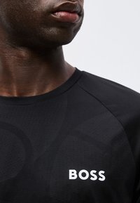 Κοντινό πλάνο ενός ατόμου που φοράει ένα μαύρο πουκάμισο με τη λέξη «BOSS» τυπωμένη με άσπρα γράμματα στο στήθος, σε ένα απλό ανοιχτό φόντο.