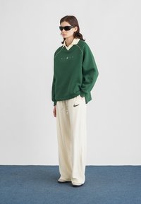 Sweatshirt verde oversize com gola amarela contrastante, calças de fato de treino creme com pequeno logótipo e óculos de sol. Texturas suaves e corte descontraído.