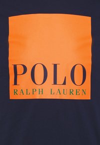 Marinblå T-shirt med en stor orange rektangel med texten "POLO" i feta mörka bokstäver ovanför "RALPH LAUREN" i mindre grön text.