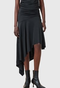Femme portant une jupe asymétrique noire avec un détail froncé à la taille et des bottines noires à fermeture éclair latérale, debout devant un fond uni.