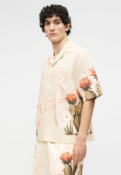 Νέος άνδρας που φοράει μπεζ πουκάμισο με κοντά μανίκια και κουμπιά καθώς και σορτς σετ, με μεγάλο πορτοκαλί σχέδιο λουλουδιού κάκτου στη αριστερή πλευρά.