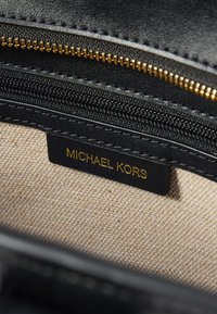 Intérieur en cuir noir avec fermeture éclair dorée et doublure beige. Comprend une étiquette noire avec "MICHAEL KORS" en texte doré.