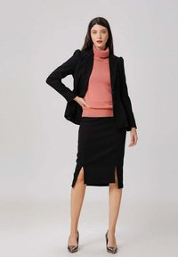 Femme portant un blazer noir, une jupe crayon fendue noire, un pull col roulé corail et des escarpins métalliques à bout pointu, se tenant avec une main sur la hanche.