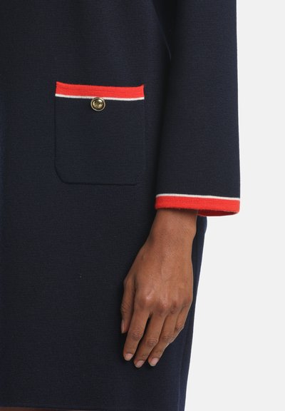 Ženská ruka vedľa námorníckeho šatstva s vreckom a lemovkou v červenej a bielej farbe, vrecko zabezpečené zlatým gombíkom.