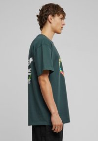Mørkegrøn bomulds t-shirt med korte ærmer. Har farverigt grafisk tryk på ryggen og logo tekst på venstre ærme. Blød tekstur.