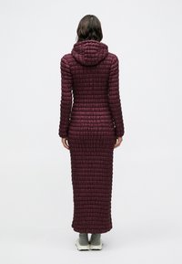 Bordeauxrote Steppjacke mit Kapuze, zeichnet sich durch ein langes, gesteppte Design und eine strukturierte Oberfläche aus. Endet am Knöchel mit einer taillierten Form.