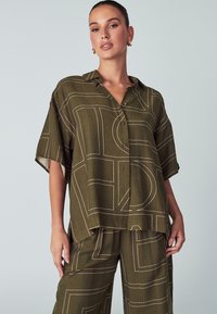 Olivgrünes Kurzarmhemd mit Kragen. Besteht aus einem geometrischen weißen Muster und hat eine Knopfleiste. Hergestellt aus einem strukturierten Stoff.