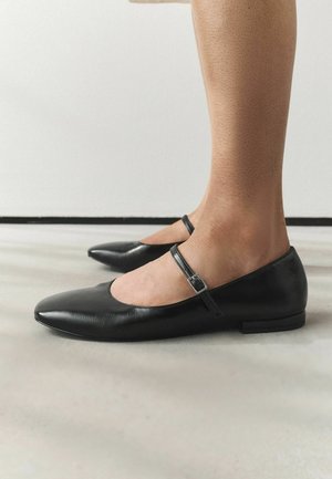 Osoba nosząca czarne skórzane baleriny Mary Jane z paskiem i sprzączką, stojąca na gładkiej jasnoszarej podłodze na tle białej ściany.