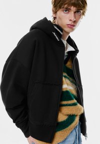 Fekete cipzáras kapucnis pulóver, laza szabással, nyers szegéllyel, puha anyagból készült, és logó található a kapucnin. Rétegezve egy csíkozott bolyhos pulóverrel.