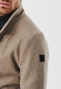 Beżowa kurtka z wełny o wysokim kołnierzu i teksturowanym wykończeniu. Posiada małą czarną naszywkę z logo na lewym ramieniu.