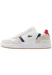 Baskets blanches avec des accents en daim beige et bleu marine, dotées de détails perforés, d'une semelle en caoutchouc et d'un capuchon de talon rouge. Logo Lacoste présent.