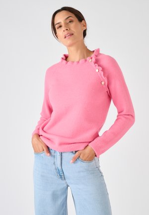 Pull rose en matière douce avec col à volants, boutons sur l'épaule et manches longues. Associé à un jean bleu clair.
