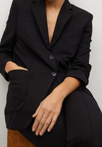 Černý blazer s klopami, dvěma knoflíky a přední kapsou; hladká látka; mírně ohrnuté rukávy; v kombinaci s odpovídajícími kalhotami.
