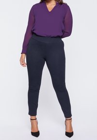 Lila genomskinlig blus med lång ärm och V-ringning, matchad med figurnära marinblå leggings. Svarta högklackade skor kompletterar outfiten.