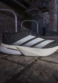 Sort og hvid Adidas løbesko med tre hvide striber og "Lightstrike Pro" på sålen, placeret på stenhøjt fortov udendørs.