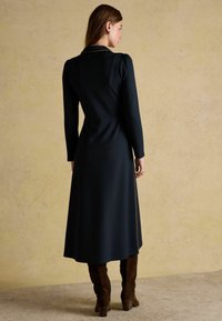 Pikk, tume sinine kleit pikkade varrukatega, kraega ja kitsas taljeosa, voolates laia seeliku poole. Kanda pruunide nahast põlvekõrguste saapadega.