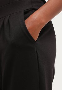 Pantaloni neri in tessuto con una superficie liscia, dotati di tasche laterali e una vestibilità sartoriale, con pieghe ordinate in vita.