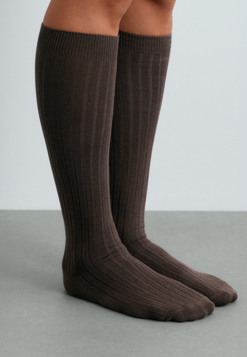 Personne portant des chaussettes côtelées brun foncé jusqu’aux genoux, debout sur une surface grise devant un fond uni.