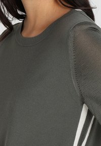 Nahaufnahme einer Person, die einen grauen Strickpullover mit durchsichtigen Netzärmeln und weißen sowie schwarzen Seitenstreifen trägt.