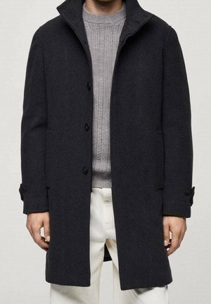 Mann trägt einen schwarzen, mit Knöpfen versehenen Wollmantel über einem grauen, gerippten Pullover und weißen Hosen, steht vor einem schlichten Hintergrund.