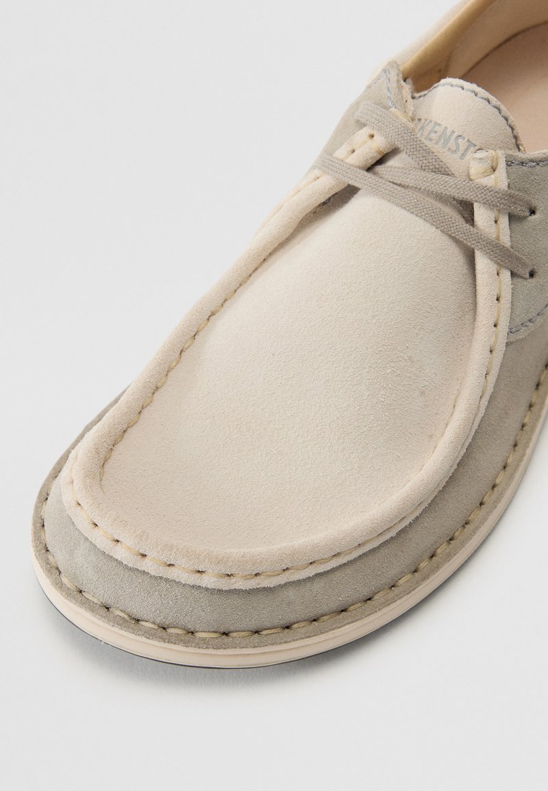 Beżowe zamszowe buty z gładką skórzaną cholewką, okrągłym noskiem i subtelnymi detalami ściegów. Posiadają jasnoszare sznurowadła i gumową podeszwę.