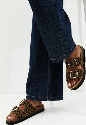 Person, die dunkelblaue Jeans und Sandalen mit Leopardenmuster sowie zwei verstellbaren goldenen Schnallen trägt, steht mit einem Fuß erhoben.