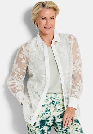 Frau mit kurzen blonden Haaren, die eine durchscheinende weiße Bluse mit Blumenmuster, ein mintgrünes Oberteil und grüne Hosen mit Blattmuster trägt, leicht lächelnd.