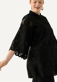 Túnica negra de manga larga con intrincados detalles de encaje y dobladillo en forma de escallona. Su cuello estructurado y las mangas acampanadas acentúan su diseño elegante.