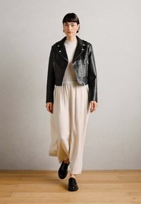 Black leather motorcycle jacket over a white, textured maxi dress with a gathered waist, paired with black shoes. Smooth, flat wooden floor.