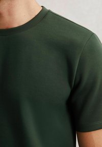 Tricou scurt cu mânecă verde închis, cu decolteu rotund, făcut dintr-un material neted și texturat. Detalii de cusătură sunt prezente pe mâneci.