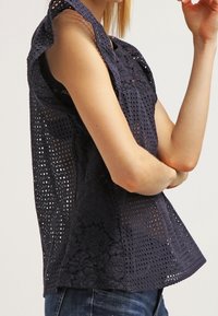 Haut sans manches bleu marine en dentelle découpée au laser avec motifs floraux et finition texturée. Caractérisé par une coupe décontractée et un ourlet arrondi.