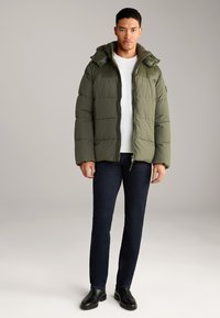 Grüne gepolsterte Jacke mit Kapuze, Reißverschluss und gesteppten Details, kombiniert mit dunklen Jeans und schwarzen Lederstiefeln.