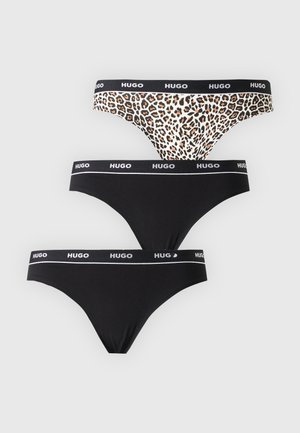 Femme aux longs cheveux foncés portant un soutien-gorge et des sous-vêtements imprimés léopard avec des bandes noires de marque Hugo, debout sur un fond uni.