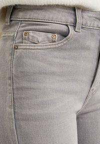 Helle graue Denim-Jeans mit klassischem Fünf-Taschen-Design, messingfarbenen Hardware-Akzenten und subtilen Texturvariationen im gesamten Stoff.