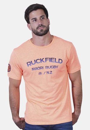 Homme aux cheveux courts et foncés et à la barbe, portant un t-shirt pêche avec le texte "RUCKFIELD MAORI RUGBY N.Z", debout les mains dans le dos.