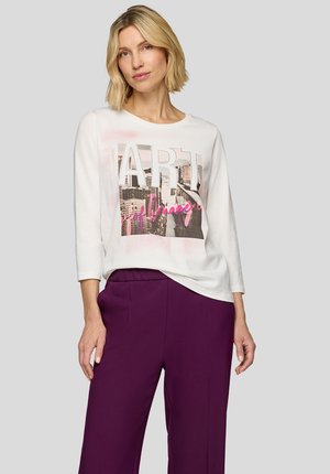 Weißes Langarmoberteil mit grafischem Druck, das das Wort "ART" und ein Stadtlandschaftsdesign zeigt. Kombiniert mit lila Hosen.