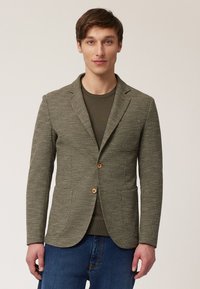 Olivengroene blazer van textuurstof, met twee knopen, ingeknepen revers en voorzakken, gedragen over een donkere top.