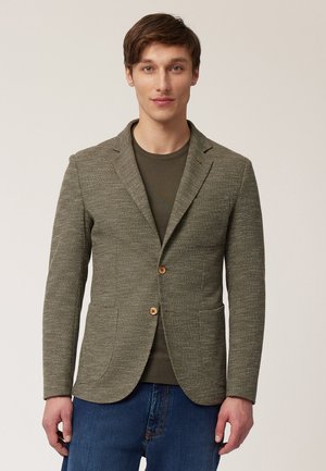 Olive green blazer made of textured fabric, featuring two buttons, notched lapels, and front pockets, worn over a dark top.